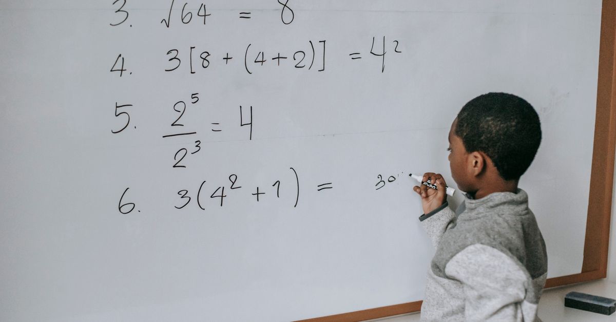 Side view of concentrated African American pupil standing near whiteboard in classroom and solving math examples during lesson at school